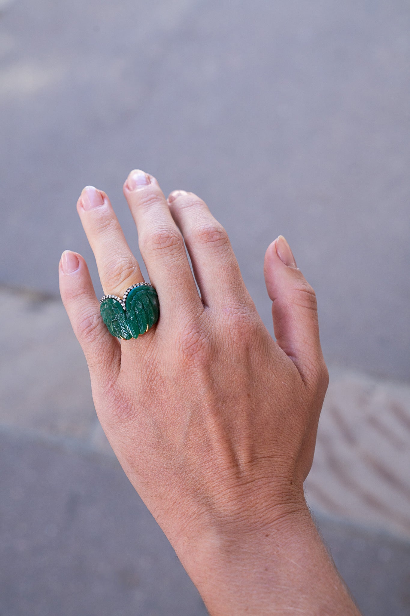 Hand wearing a green ring against a blurred background. Carved-Green-Dumortierite-Feather-Ring-Silvia-Furmanovich-510466995
