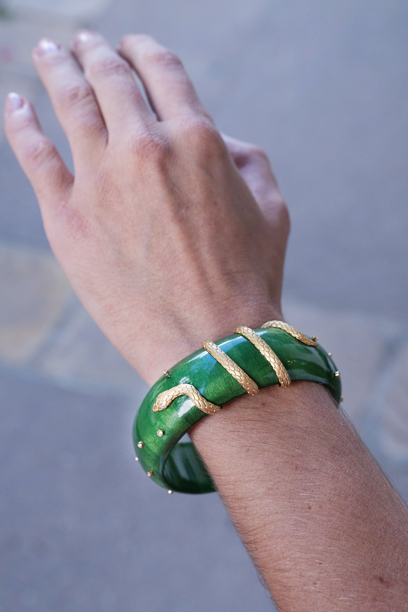 Hand wearing a green bracelet with gold accents on a blurred background. Green-Lacquered-Marquetry-Bangle-with-Snake-Detail_-Silvia-Furmanovich-