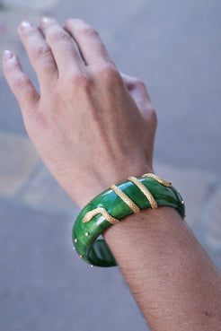 Hand wearing a green bracelet with gold accents on a blurred background. Green-Lacquered-Marquetry-Bangle-with-Snake-Detail_-Silvia-Furmanovich-