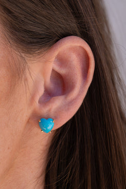 Close-up of an ear wearing a blue heart-shaped earring with a blurred background. Irene Neuwirth Heart Turquoise Studs