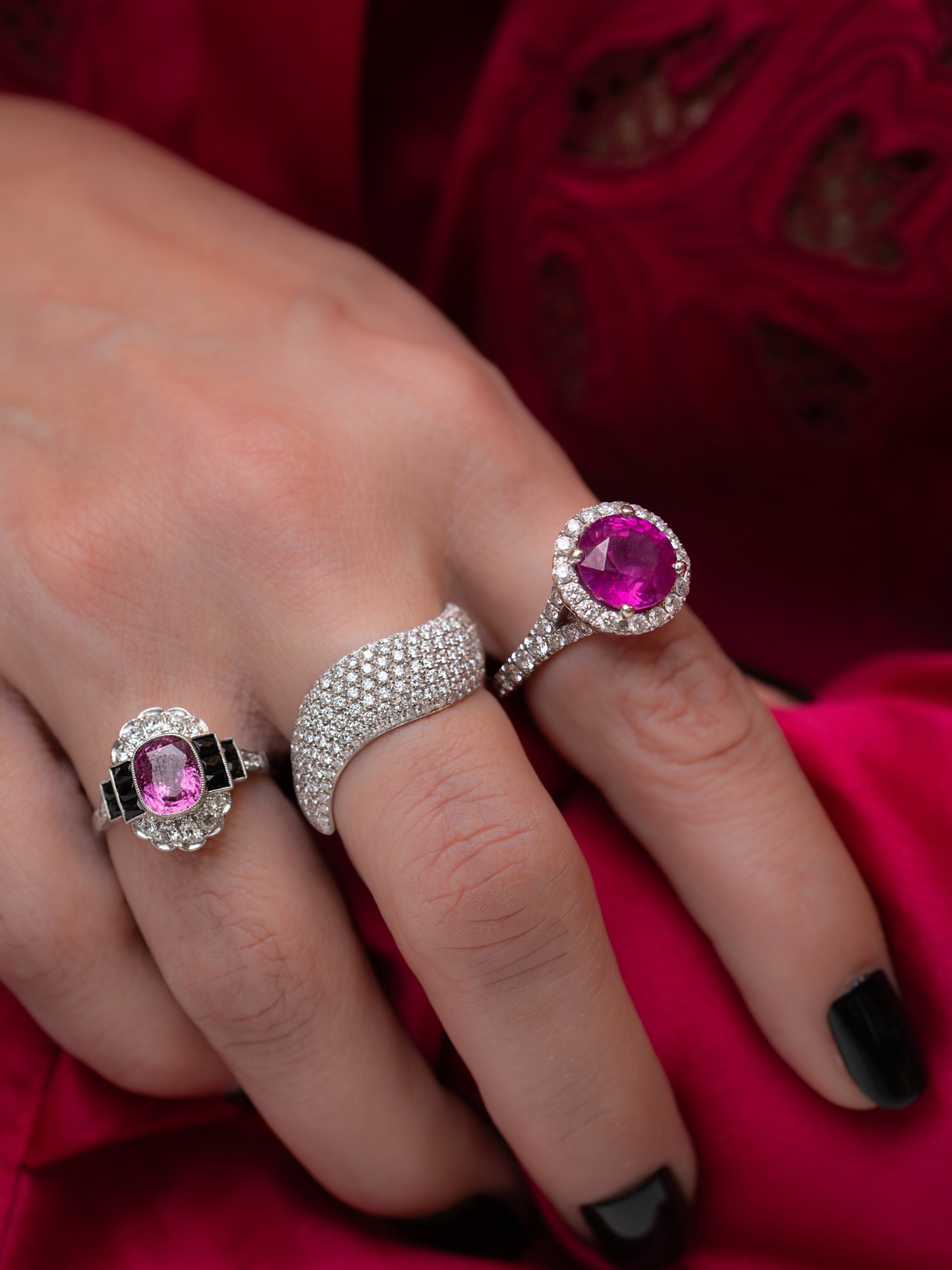 Three rings displayed on a hand, including a silver ring with a pink gemstone, a silver pavé band ring, and a silver halo ring with a larger pink gemstone, set against a red fabric background.