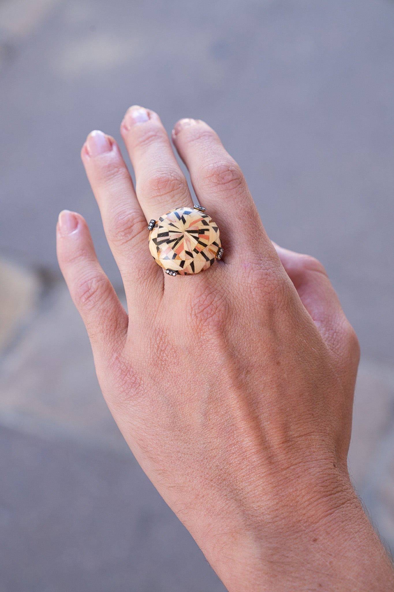 Hand wearing a large ring with a textured design on a blurred background. Silva_Furmanovich_Maquetry_Ring